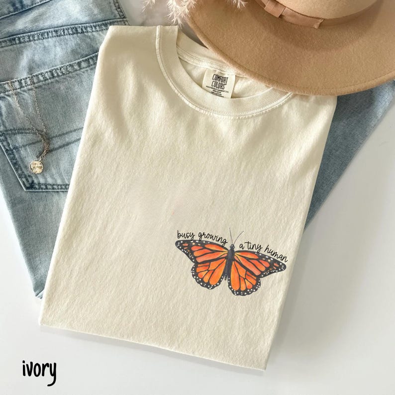 May include: Ivory-colored t-shirt with a butterfly graphic and the text "busy growing a tiny human." The shirt is laid on top of blue jeans and a tan hat.