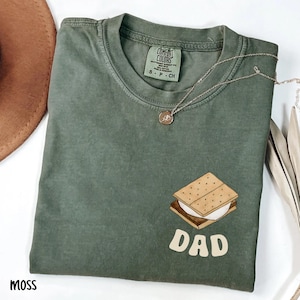 May include: A moss green t-shirt with a s'mores graphic and the word "DAD" in a retro font. A silver necklace with a pendant is draped across the shirt. The shirt is folded and has a tag that says "Comfort Colors".