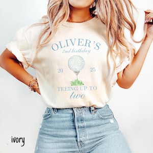 May include: A light beige t-shirt with a blue and white golf ball graphic and the text "Oliver's 2nd birthday Teeing Up To Two" printed on it.