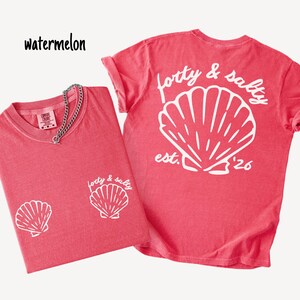 May include: Two watermelon-colored t-shirts with white seashell graphics. One shirt has a front graphic and a chain necklace. The other shirt has a large seashell graphic on the back with the text "forty & salty est. '26". The word "watermelon" is written in black.