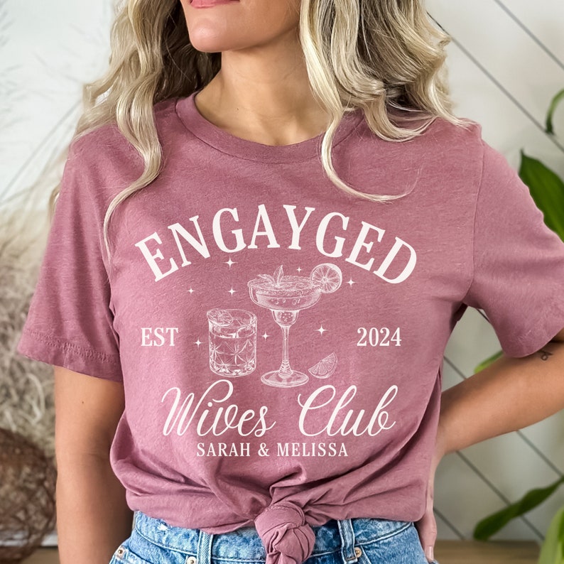 May include: A pink t-shirt with the text "Engayged Wives Club" in white. The text is surrounded by two cocktail glasses and the year 2024. The text "EST" is to the left of the glasses and the names "Sarah & Melissa" are below the glasses.