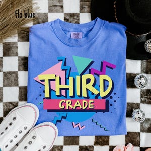 May include: A blue t-shirt with the words "THIRD GRADE" in yellow, pink, and purple, with a retro geometric design. The shirt is paired with white sneakers. The words "Flo blue" are in the top left corner.
