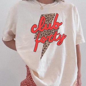 May include: Cream-colored t-shirt with the text "Club Forty" in red script over a leopard print lightning bolt design. The shirt has short sleeves and a crew neck.