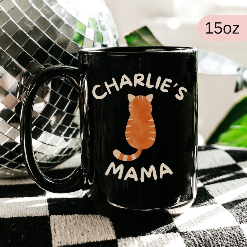 May include: Black 15oz ceramic mug with the words "Charlie's Mama" in white. An orange tabby cat illustration is centered between the words. The mug has a large black handle.