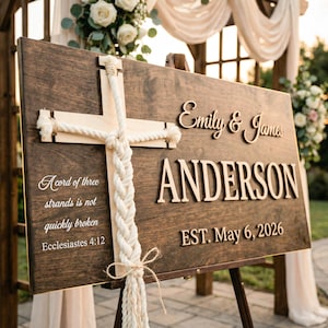 May include: A dark stained wooden wedding sign with a cross made of light wood and rope. The sign includes the names "Emily & James" and "ANDERSON", with the date "EST. May 6, 2026". A quote from Ecclesiastes is also featured.