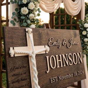 May include: A wooden wedding sign with the names "Emily & James JOHNSON" and the date "EST. November 18, 2024". A light-colored wooden cross is attached with rope. The sign includes the quote "A cord of three strands is not quickly broken. Ecclesiastes 4:12".