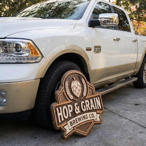 May include: A wooden sign featuring a carved hop cone and wheat stalks above the text HOP & GRAIN BREWING CO. The sign has a layered, multi-dimensional design with dark brown stained wood accents and a light wood finish on the lettering and border.