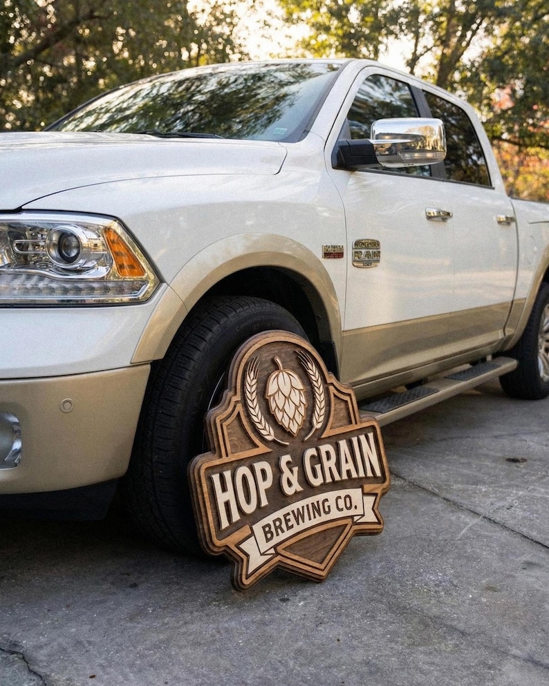 May include: A wooden sign featuring a carved hop cone and wheat stalks above the text HOP & GRAIN BREWING CO. The sign has a layered, multi-dimensional design with dark brown stained wood accents and a light wood finish on the lettering and border.