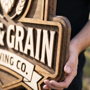 May include: A multi-layered wooden sign featuring a dark brown stained finish with light natural wood accents. The sign displays the text GRAIN and WING CO. along with a hop cone graphic. The design has a raised, dimensional structure.