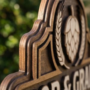 May include: A layered wooden sign with a dark brown stained finish featuring a carved hop cone and wheat stalks. The text HOP & GRAIN is prominently displayed in raised, light-coloured wood lettering against the darker, multi-tiered wooden background.