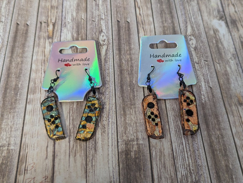 May include: Two pairs of handmade earrings featuring a Nintendo Switch Joy-Con controller design. The earrings are made with a gold and black glitter finish and have black hooks. The earrings are displayed on a white card with the text "Handmade with love".
