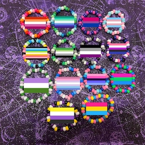 May include: A collection of 12 colorful beaded bracelets, each featuring a different pride flag design. The bracelets are arranged in a circle on a purple background with a starry pattern.