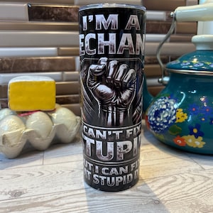 Pode incluir: Um copo preto com o texto "I'M A MECHANIC, CAN'T FIX STUPID, BUT I CAN FIX STUPID". O copo apresenta um gráfico de um punho cerrado. O copo é cilíndrico e parece ser feito de aço inoxidável.