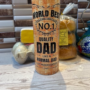 Pode incluir: Um copo cilíndrico com um design de grão de madeira. O copo apresenta o texto "WORLD BEST NO.1 QUALITY DAD LIKE A NORMAL DAD JUST MUCH COOLER" em letras pretas. O fundo inclui produtos de padaria e uma parede de azulejos decorativos.