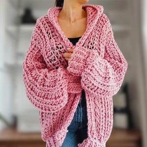 Op de afbeelding: Een roze gebreide cardigan met een capuchon en oversized mouwen. De cardigan is gemaakt van een dik garen en heeft een losse, ontspannen pasvorm.