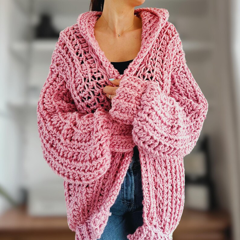 Op de afbeelding: Een roze gebreide cardigan met een capuchon en oversized mouwen. De cardigan is gemaakt van een dik garen en heeft een losse, ontspannen pasvorm.