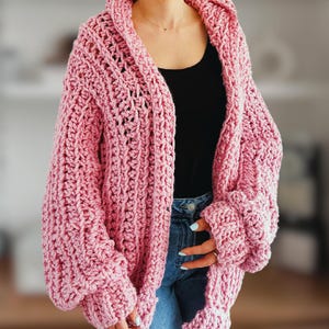Op de afbeelding: Een roze gebreide cardigan met een losse pasvorm en lange mouwen. De cardigan heeft een V-hals en een capuchon.