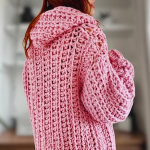 Puede incluir: Un cárdigan de color rosa tejido a crochet con capucha y mangas largas. El cárdigan tiene un corte holgado y de gran tamaño.