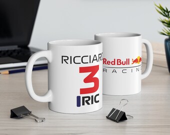 May include: Two white ceramic mugs with a Formula One racing theme. The mug on the left has the number 3 and the name Ricciardo with the initials RIC. The mug on the right has the Red Bull Racing logo.
