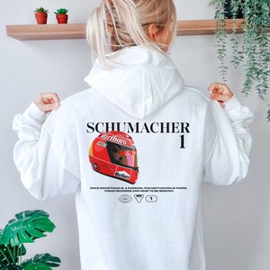 Pu&ograve; includere: Felpa bianca con un grafico di casco da corsa rosso e bianco, il testo "SCHUMACHER 1" e il testo "ONCE SOMETHING IS A PASSION, THE MOTIVATION IS THERE. THESE RECORDS ARE HERE TO BE BROKEN."