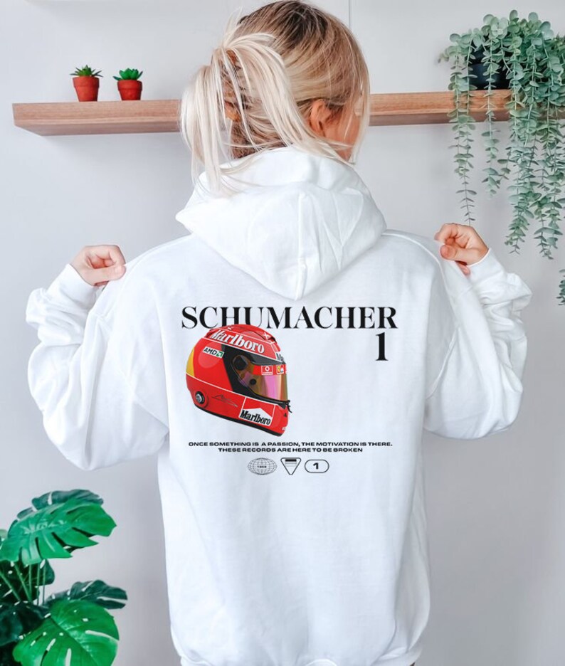 Pu&ograve; includere: Felpa bianca con un grafico di casco da corsa rosso e bianco, il testo "SCHUMACHER 1" e il testo "ONCE SOMETHING IS A PASSION, THE MOTIVATION IS THERE. THESE RECORDS ARE HERE TO BE BROKEN."