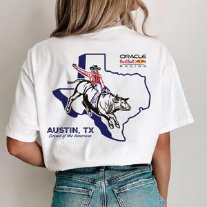 以下が含まれることがあります： 白いTシャツに、テキサス州の青と白の輪郭が描かれています。輪郭の中に、漫画の牛仔が牛に乗っています。「Oracle Red Bull Racing」と「Austin, TX Circuit of the Americas」の文字がTシャツに印刷されています。
