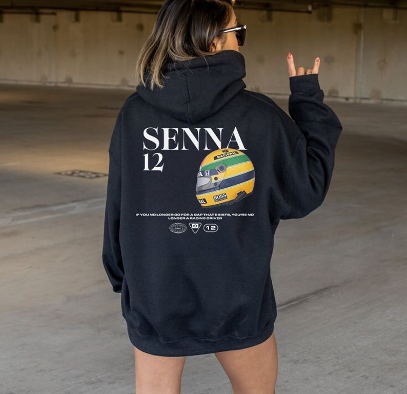 Puede incluir: Sudadera con capucha negra con un estampado gr&aacute;fico blanco de un casco de carreras y el texto "SENNA 12" y la cita "Si ya no vas por un hueco que existe, ya no eres un piloto de carreras".
