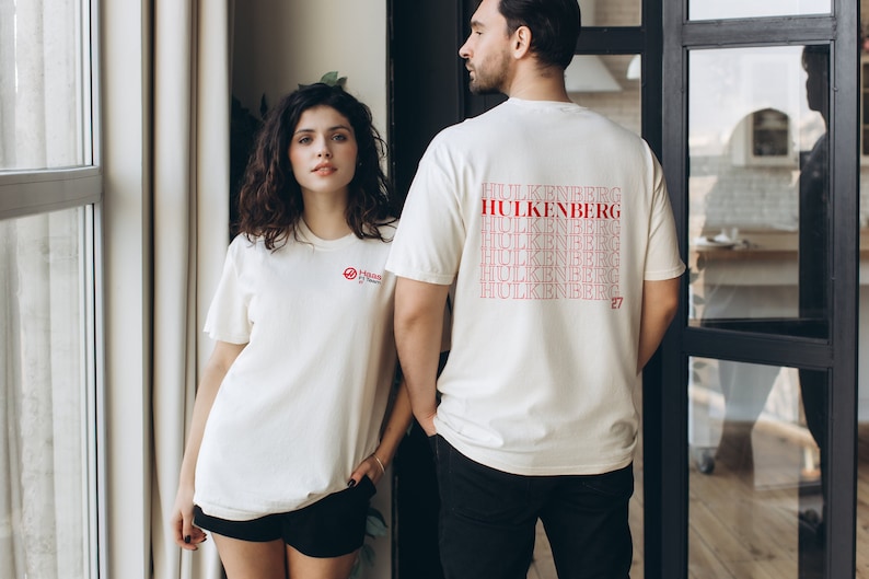 May include: Two people wearing white t-shirts. The person on the right is wearing a t-shirt with the text "HULKENBERG" repeated multiple times in red. The number "27" is printed in red on the bottom right of the shirt.