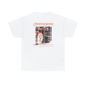 May include: White t-shirt with a red and white image of Ayrton Senna in a race car. The text "Ayrton Senna" and "1960" are printed on the shirt.