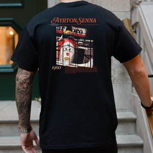 May include: Black t-shirt with a graphic print of a race car driver in a red and white race car. The text "Ayrton Senna 1960" is printed on the shirt.