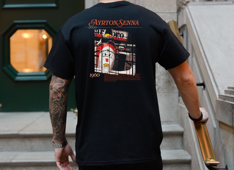 May include: Black t-shirt with a graphic print of a race car driver in a red and white race car. The text "Ayrton Senna 1960" is printed on the shirt.