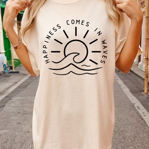 May include: A cream colored t-shirt with a black graphic design of a sun and wave. The text on the shirt reads "Happiness comes in waves".
