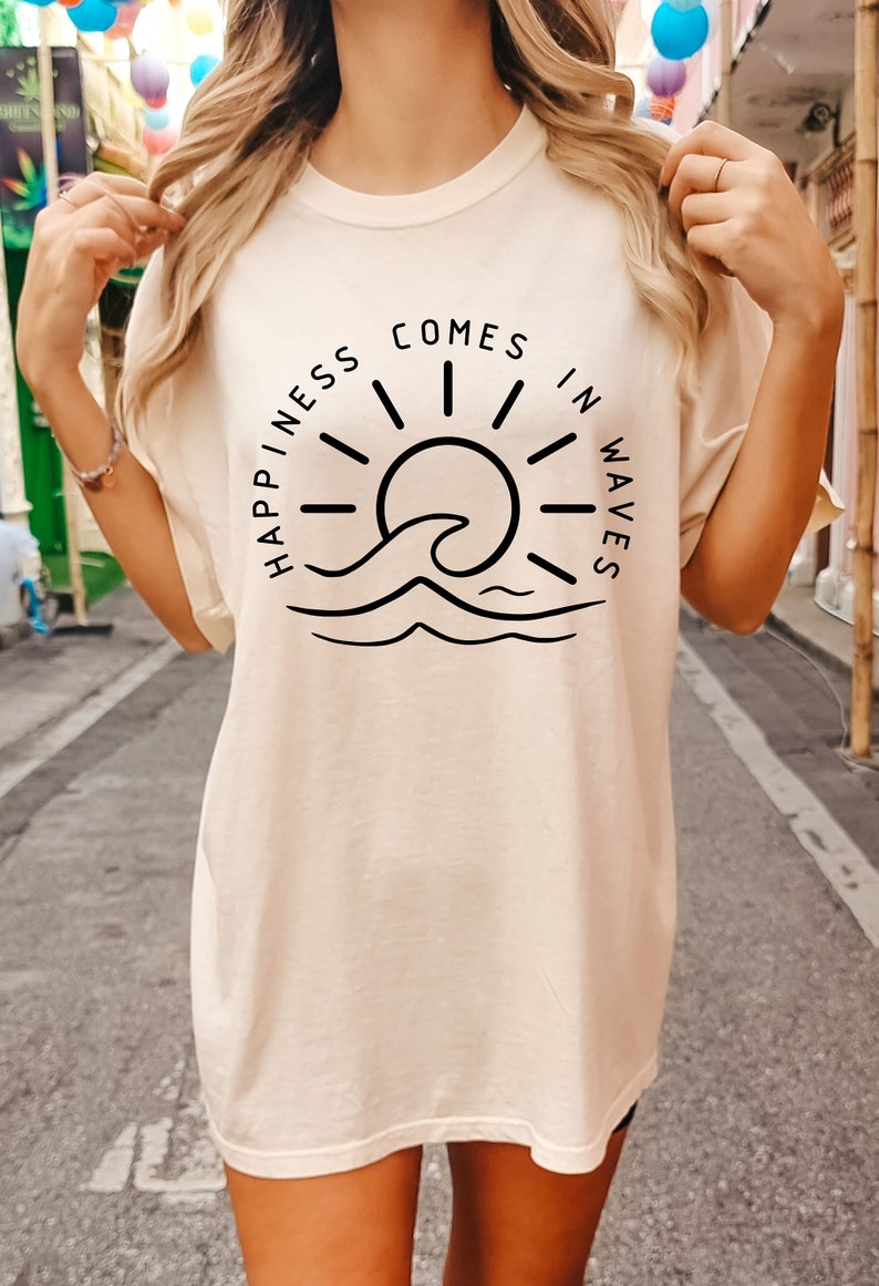 May include: A cream colored t-shirt with a black graphic design of a sun and wave. The text on the shirt reads "Happiness comes in waves".
