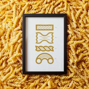 May include: A black framed print with a white background and gold illustrations of different types of pasta. The illustrations include a long noodle, a bowtie pasta, a twisted noodle, and a curved noodle.
