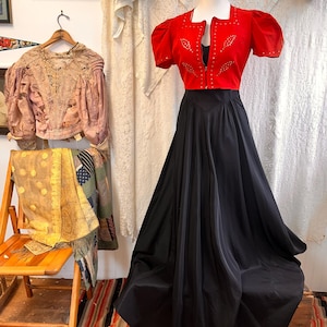 May include: A vintage dress displayed on a mannequin. The dress comprises a red, short-sleeved top with embellishments and a black, flowing skirt. The top has a square neckline and a zip. The skirt is long and full, creating a classic silhouette.