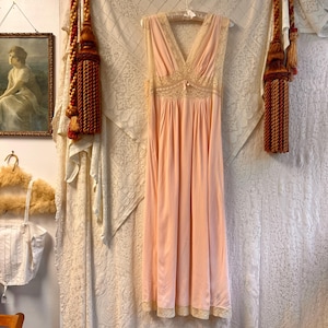 May include: A sleeveless floor-length gown in a soft peach color featuring a deep V-neckline, a fitted bodice with intricate cream-colored lace detailing, a small central bow, and a flowing skirt that ends with a matching lace trim at the hem.
