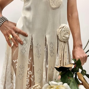 May include: Ivory-colored vintage dress with a lace overlay and floral embellishments. The dress features intricate beading and a flowing design. A bouquet of white roses is held in the hand.