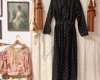 Puede incluir: Un vestido negro vintage con lunares blancos, mangas largas y cintura con cinturón. El vestido está colgado de una percha de madera. El vestido es de estilo clásico, adecuado para una variedad de ocasiones.