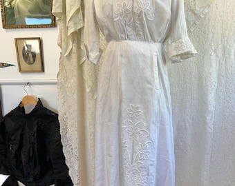 Puede incluir: Un vestido blanco de algodón con delicados bordados florales. El vestido tiene un corpiño ajustado y una falda fluida. El bordado está hecho con hilo blanco y es muy intrincado.