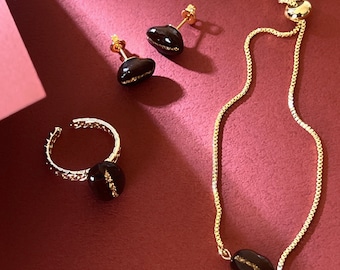 May include: A jewelry set featuring a ring, earrings, and a bracelet, all with a coffee bean theme. The gold-toned pieces have dark brown coffee bean-shaped accents with gold detailing. The text "KONA COFFEE" is visible.