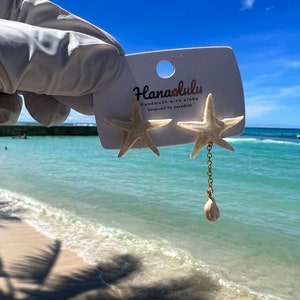 Pu&ograve; includere: Orecchini a forma di stella marina con un ciondolo a forma di conchiglia. Gli orecchini sono attaccati a una carta bianca con il testo "Hanaolulu Handmade with aloha Inspired by paradise". Lo sfondo &egrave; una spiaggia con cielo blu e acqua.