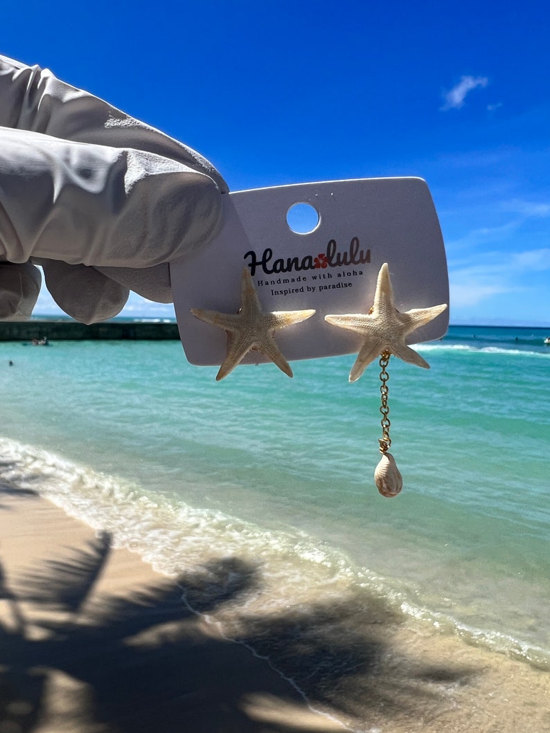Pu&ograve; includere: Orecchini a forma di stella marina con un ciondolo a forma di conchiglia. Gli orecchini sono attaccati a una carta bianca con il testo "Hanaolulu Handmade with aloha Inspired by paradise". Lo sfondo &egrave; una spiaggia con cielo blu e acqua.