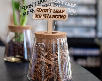 Puede incluir: Un tarro de vidrio con tapa de corcho y virutas de papel marrón en su interior. Un palo de madera con un letrero que dice "Don't Leaf Me Hanging" está insertado en el corcho.