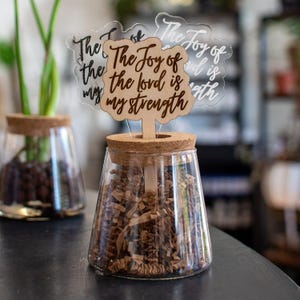 May include: A glass jar with a cork lid and a wooden stick with a sign that reads "The Joy of the Lord is my Strength". The jar is filled with brown paper shreds and a green plant is growing in the background.