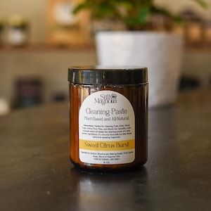May include: A brown glass jar with a black lid containing cleaning paste. The label reads "Still Magnolia Cleaning Paste Plant-Based and All-Natural Sweet Citrus Burst" and lists the ingredients as "Sodium Bicarbonate, Baking Soda, Pure Castile Soap, Blend of Essential Oils". The jar is 8 oz and for external use only.