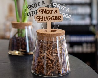 Puede incluir: Un tarro de cristal transparente con tapa de corcho y la frase "Not a Hugger" en un letrero de madera. El tarro está lleno de papel triturado marrón. Otro tarro con una planta está en el fondo.