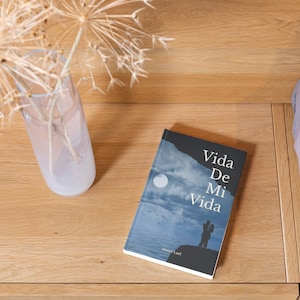 May include: A book titled "Vida De Mi Vida" by Anaya Leal with a blue and white cover featuring a silhouette of a couple standing on a cliff overlooking a moonlit ocean.