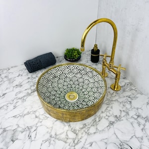 Puede incluir: Un lavabo de baño de cerámica blanco y dorado con un diseño floral verde. El lavabo es redondo y tiene un borde dorado. Un grifo dorado está montado en la pared sobre el lavabo.