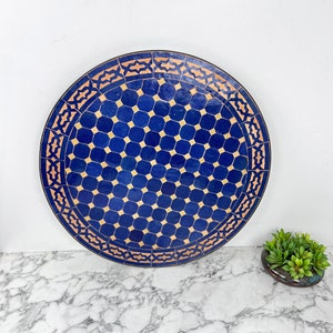 Puede incluir: Una bandeja redonda de azulejos marroquíes azul y crema con un patrón geométrico. La bandeja tiene un borde de color cobre.