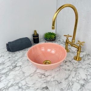 Peut inclure: Un lavabo de salle de bain en céramique rose avec un robinet doré. Le lavabo est posé sur un plan de travail en marbre blanc.